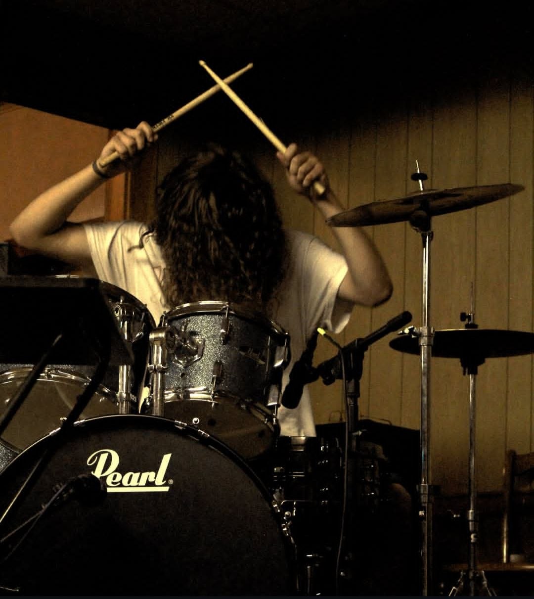 Sepia-toned drummer from behind, sticks crossed overhead
