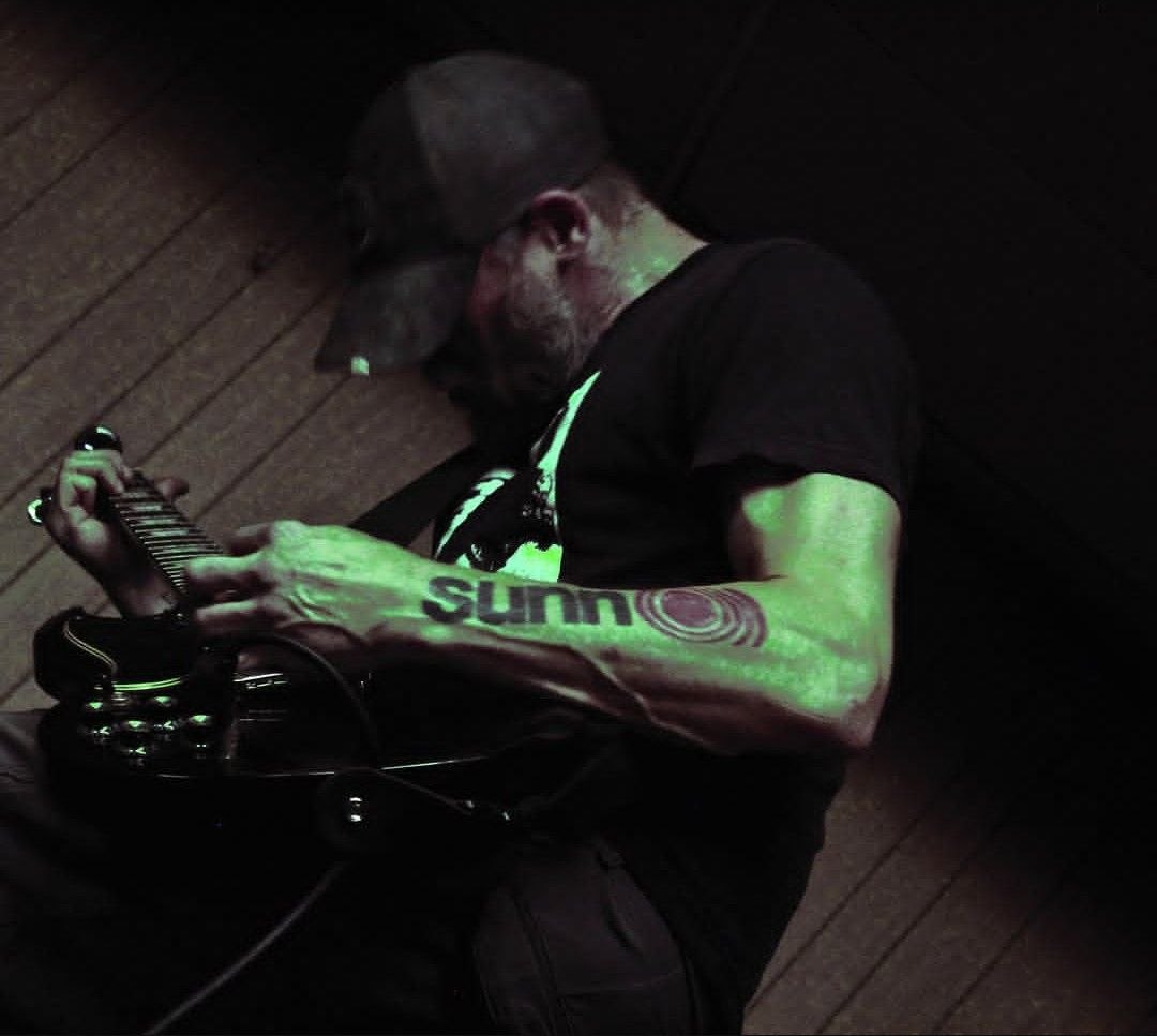 Guitarist under green stage light, head down, Sunn tattoo visible on his arm