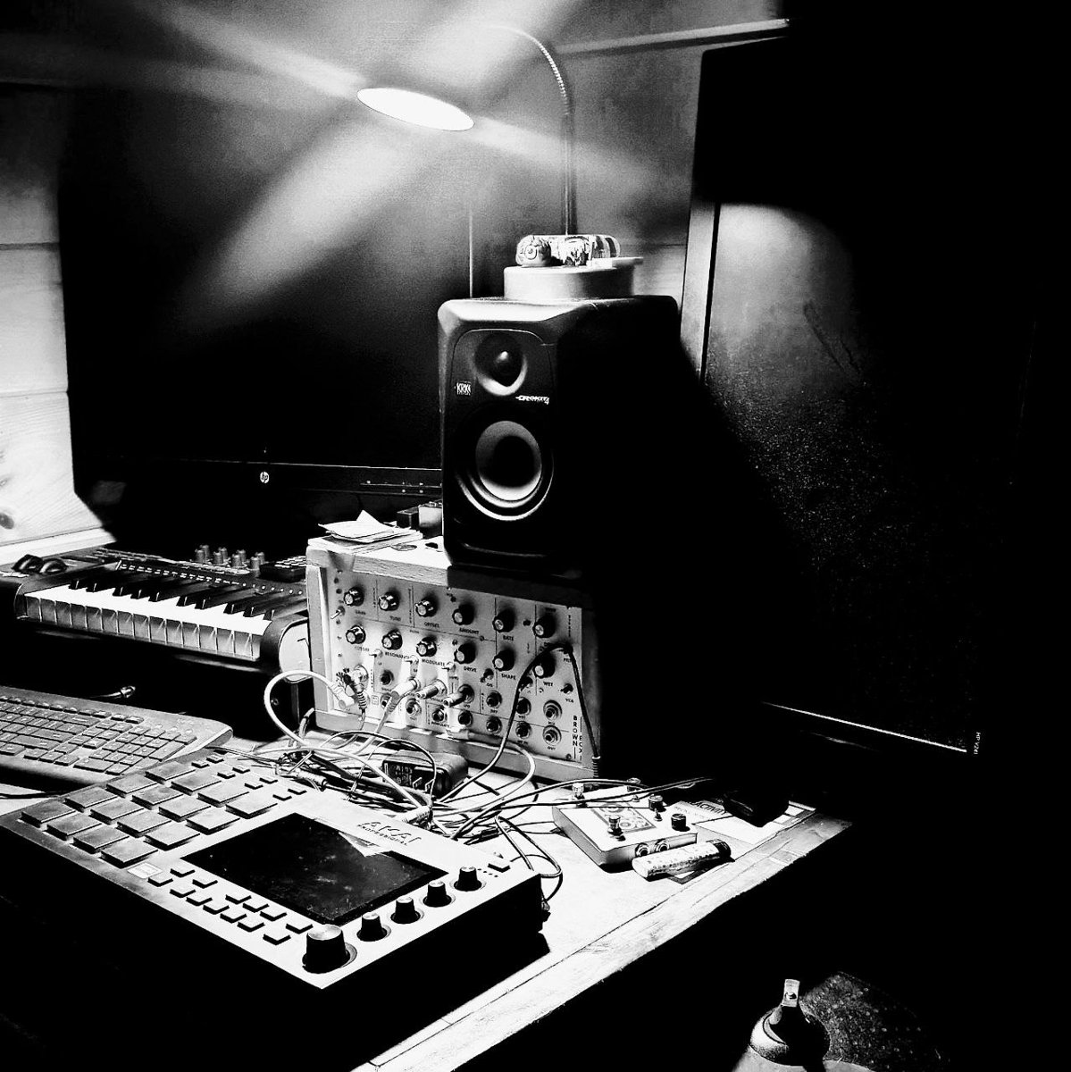 Black and white photo of the home studio: synth modules, an MPC, keyboard, and a monitor speaker under a work lamp